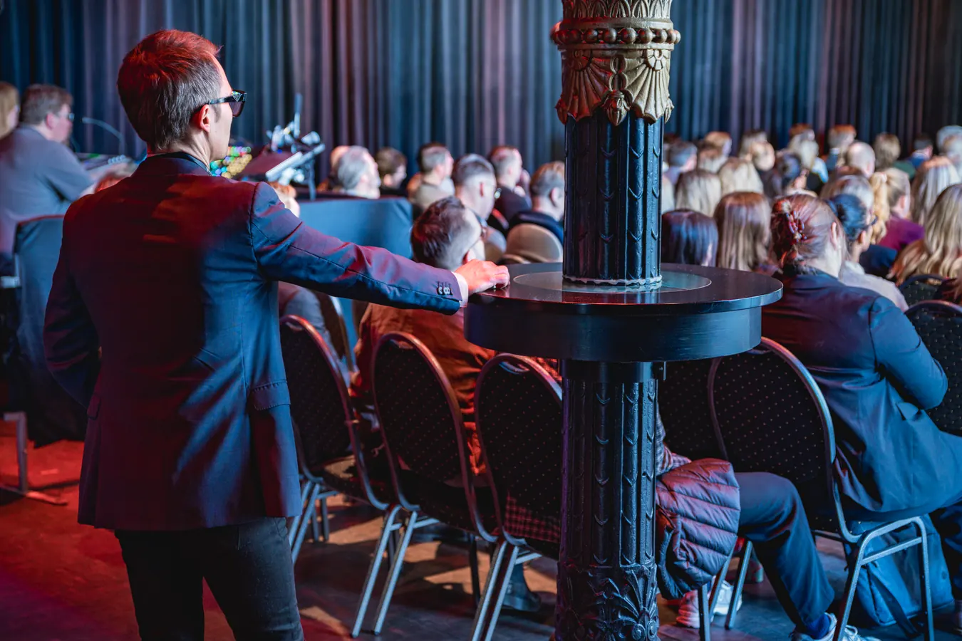 Man in a suit seen from behind, hand resting on a pedestal table beside an ornate pillar at a conference